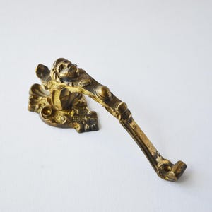 Könnte beinhalten: Ein goldfarbener Metallhaken mit einem verzierten Design mit einem Gesicht und Schnörkeln. Der Haken ist für die Wandmontage konzipiert.