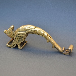 Pode incluir: Uma maçaneta decorativa de cor latão em forma de pássaro. O pássaro tem asas detalhadas e um corpo curvo, com uma extremidade enrolada. A maçaneta é projetada para ser montada em uma porta.