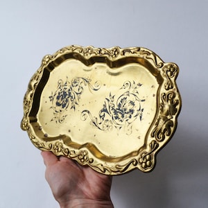Puede incluir: Una bandeja vintage de metal dorado con un borde festoneado adornado. La bandeja presenta un diseño floral en azul oscuro. La bandeja está sujeta por una mano. La bandeja probablemente se utiliza para servir o exhibir.