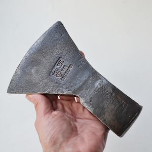 Peut inclure: Une tête de hache en métal gris foncé avec une lame large et incurvée et un talon rectangulaire. La surface présente une marque estampée contenant le texte 800, un symbole triangulaire et les lettres TGL GDR, indiquant le poids et l'origine.