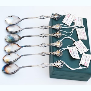May include: A set of six silver-toned metal spoons with ornate, openwork handles featuring a geometric lattice design. Each spoon is attached to a small white paper tag with handwritten text and rests beside a rectangular dark green storage box.