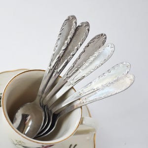 Può includere: Un set di quattro cucchiai in metallo argentato con motivi a volute in rilievo sui manici, disposti a ventaglio all'interno di una tazza da tè in ceramica color crema con un delicato disegno floreale sulla parte esterna.