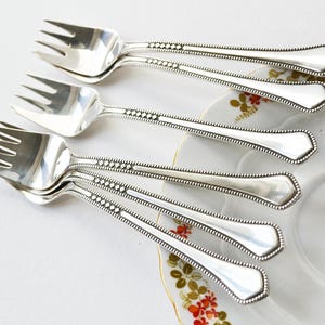 May include: A collection of silver forks with three prongs, displayed on a white plate with a floral pattern. The forks feature a beaded border and a geometric design on the handle. The cutlery is reflective and looks vintage.