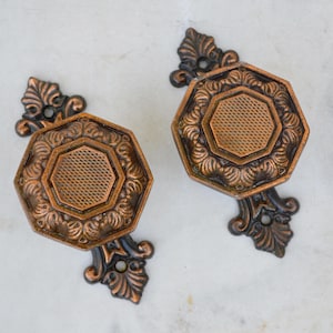 May include: Two ornate copper door knobs with intricate floral designs and a black metal base. The knobs are octagonal in shape and have a textured surface.