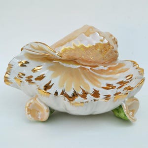May include: A ceramic decorative piece in the shape of a flower, with a pearlescent finish. The petals are white with gold accents and a peach-colored overlay. Green accents are visible at the base.