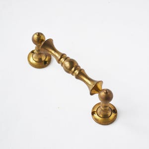 Puede incluir: Un tirador de puerta de latón antiguo con un diseño decorativo. El tirador presenta una sección central alargada con detalles redondeados y placas de montaje circulares. El latón tiene una pátina cálida y envejecida.