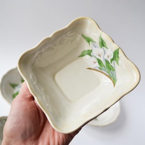 May include: A square, off-white ceramic bowl with scalloped edges and a raised scroll pattern along the rim. The interior features a painted illustration of white calla lilies with green leaves extending from the corner toward the center.