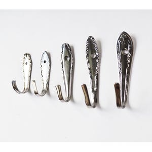 May include: Five silver metal wall hooks made from repurposed silverware. The hooks are arranged in a row, each with a different size and design. The hooks are mounted on a white wall, showcasing their unique, vintage aesthetic.