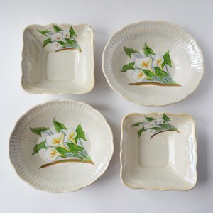May include: Four off-white ceramic bowls with scalloped edges. Each bowl features a hand-painted design of white calla lilies with yellow centers and green leaves. The bowls are in both square and round shapes.