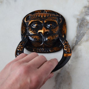 以下が含まれることがあります： ライオンの顔が特徴的な、黒と金の装飾的なドアノッカー。ノッカーは、リング状のハンドルを備えた詳細なデザインです。ライオンの顔は金色の鼻と口をしています。ノッカーは白い表面に取り付けられています。