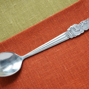 Peut inclure: Une cuillère en argent avec un motif d'ours sur le manche, posée sur un tissu orange. La cuillère a un bol arrondi et une figurine d'ours détaillée en haut. Le fond est un tissu vert contrastant.
