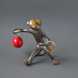 May include: A whimsical, metal monkey figurine with a red sphere earring and a gold ring with turquoise accents. The monkey has a distressed, aged appearance with gold accents on its face and feet. The figurine is a unique jewelry holder.