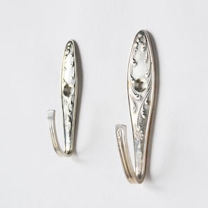 Può includere: Due ganci da parete color argento realizzati con posate riciclate. I ganci presentano un motivo floreale decorativo e una forma a gancio ricurva. Sono montati su una parete bianca, mettendo in risalto il loro design elegante e il potenziale per l'arredamento.