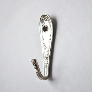May include: A silver-toned metal wall hook crafted from a repurposed spoon handle. The hook features an elongated, flat backplate with intricate embossed scrollwork patterns and a single circular mounting hole near the top, curving upward.