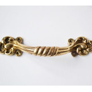 May include: A gold-toned metal drawer pull featuring a central rope-textured section flanked by ornate, curved scrollwork ends. The handle includes two circular mounting holes on each side, showcasing a polished finish with intricate detailing.
