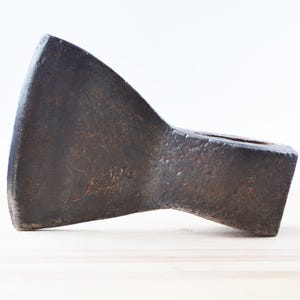Könnte beinhalten: Ein antiker Axtkopf mit dunklem, verwittertem Aussehen. Das Metallwerkzeug hat eine gebogene Klinge und ein rechteckiges Auge für den Stiel. Der Axtkopf hat eine dunkelbraune, rostfarbene Farbe.