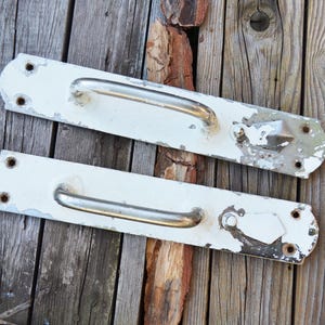 Może przedstawiać: Dwa białe, zabytkowe uchwyty do drzwi ze srebrnymi uchwytami. Prostokątne uchwyty mają zaokrąglone końce i noszą ślady zużycia, w tym odpryski farby. Tło to zwietrzałe drewno.