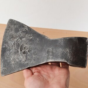 Peut inclure: Une tête de hache en métal gris foncé avec un tranchant large et incurvé, présentant une surface patinée et texturée. Le métal comporte une marque circulaire estampée près du centre de la lame, révélant un emblème avec des détails.