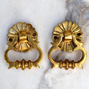 May include: Two matching gold-toned metal drawer pulls featuring a circular, fan-shaped mounting plate with two screw holes and a hinged, drop-style handle with a decorative, ridged bottom edge. The hardware has a polished, reflective finish.