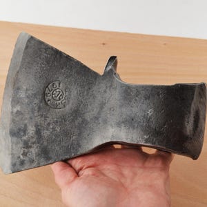 Puede incluir: Una cabeza de hacha de metal gris oscuro con una superficie texturizada y desgastada. La hoja presenta una marca circular estampada con el texto 1979 y las letras AB. La cabeza del hacha tiene un filo ancho y curvo.