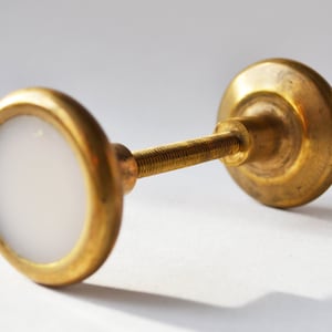 May include: A brass cabinet knob with a white center. The knob has a round face with a white inset and a threaded post for attachment. The other end of the knob is a larger, solid brass disc.