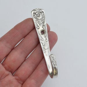 Puede incluir: Un gancho plateado hecho de una cuchara reutilizada. El gancho presenta detalles ornamentados de flores y volutas. El gancho mide aproximadamente 13 cm de largo y tiene un extremo curvado para colgar objetos. Se perfora un agujero para el montaje.