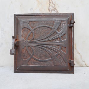 Könnte beinhalten: Quadratische, antike Gusstür mit einem verzierten Jugendstil-Design. Die Tür hat eine rostbraune Farbe und weist ein zentrales Blumenmotiv mit geschwungenen Linien und einem strukturierten Hintergrund auf. Sie hat einen Griff auf der rechten Seite und einen Riegel auf der linken Seite.