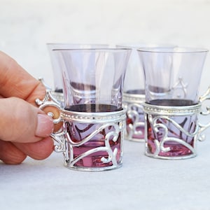 May include: A set of small purple glass cups, each encased in a decorative silver-toned metal holder with an ornate, swirling handle. The translucent purple glass contrasts with the metallic silver frame, creating a distinct visual pairing for each cup.
