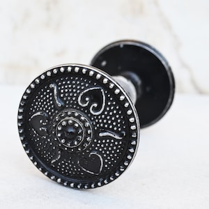 Könnte beinhalten: Schwarzer Türknauf aus Metall mit einem dekorativen silbernen Design. Der Knauf ist rund mit einer erhabenen Mitte und einem perlenbesetzten Rand.