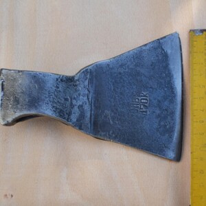 Könnte beinhalten: Ein dunkelgrauer Axtkopf aus Metall mit einer breiten, geschwungenen Schneide und einem rechteckigen Hammerende. Die Metalloberfläche hat ein texturiertes, geschmiedetes Finish mit der Prägung "JNB 470K". Ein gelbes Maßband zeigt 10 cm.