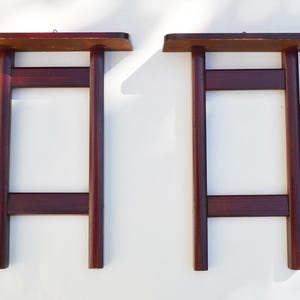 Op de afbeelding: Een paar donkerbruine houten wandplanken, elk met een plat rechthoekig bovenoppervlak ondersteund door twee verticale cilindrische poten verbonden door twee horizontale dwarsbalken, wat een stevige frameconstructie voor wandmontage vormt.