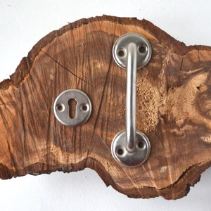 Puede incluir: Un juego rústico de manija de puerta y placa de ojo de cerradura sobre un trozo de madera natural. La manija es un tirador de metal cepillado con una barra curva. La placa del ojo de cerradura es redonda.