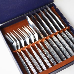 May include: A set of stainless steel flatware consisting of four forks and four knives arranged in a blue presentation box. Each handle features an intricate embossed floral pattern, and the utensils are secured with brown ribbon ties.