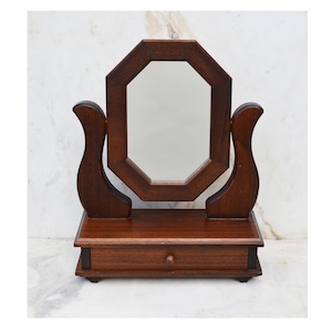 May include: A dark brown wooden vanity mirror with an octagonal mirror and a small drawer. The mirror is framed in a dark wood and has a beveled edge. The drawer has a small knob.