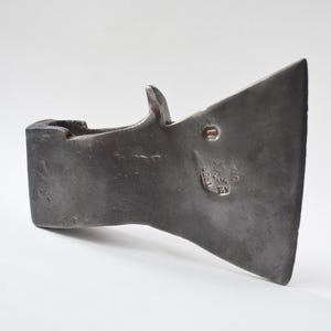 May include: A dark gray axe head with a curved blade and a rectangular eye for the handle. The axe head has a small, raised hook on the top and a circular marking with text. The metal has a worn, textured appearance.