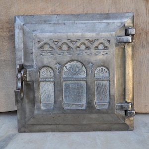 May include: A square, silver-toned metal stove door featuring a decorative arched border at the top and three vertical arched panels. The central panel displays embossed floral motifs and faint, partially obscured text. It has a side latch and hinges.