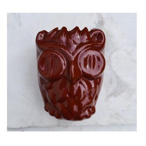 May include: A glossy, dark brown ceramic wall-mounted object shaped like an owl head. The piece features large, circular, protruding eyes, a pointed beak, and a textured surface that mimics feathers, with a scalloped top edge.