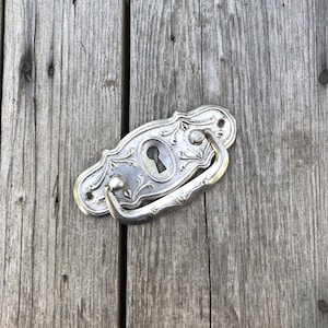 May include: A silver-coloured metal drawer pull featuring an ornate, embossed floral pattern and a central keyhole escutcheon. The hardware has a curved, swinging handle attached to a decorative backplate with two visible screw holes for mounting.