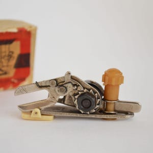 Könnte beinhalten: Nahaufnahme eines Vintage-Dosenöffners aus Metall mit beigem Kunststoffgriff und -knopf. Der Dosenöffner hat einen komplexen Getriebemechanismus und einen Hebelarm. Im Hintergrund ist eine teilweise sichtbare Pappschachtel zu sehen.