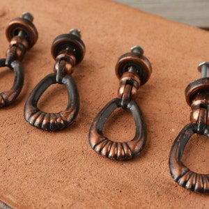 Pode incluir: Quatro botões de armário de estilo antigo com acabamento em cobre e um anel preto em torno da abertura. Os botões têm uma forma oval simples e são projetados para uso em gavetas ou portas.