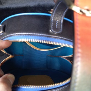 May include: A black, circular handbag with a blue interior and a blue zip. The bag features a handle and a colourful strap. The interior is split into two compartments. The bag is made of leather.