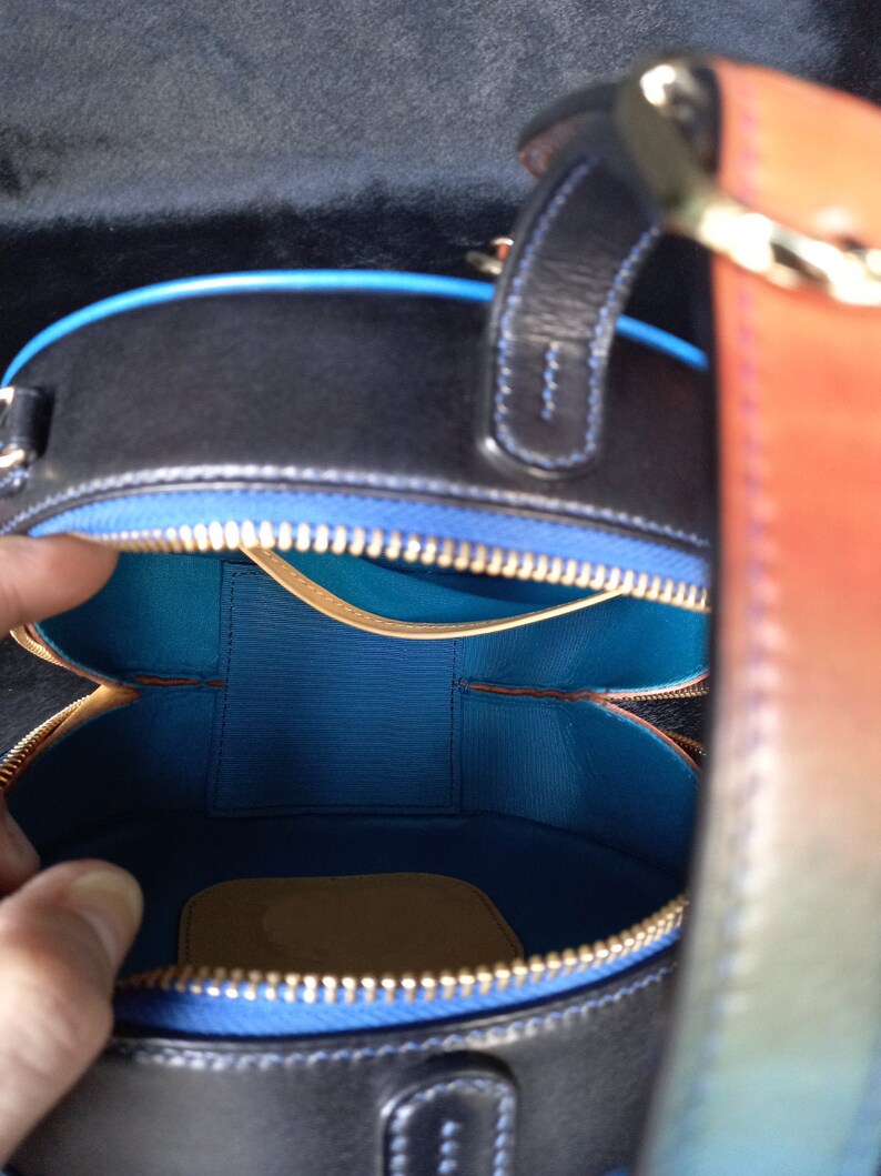 May include: A black, circular handbag with a blue interior and a blue zip. The bag features a handle and a colourful strap. The interior is split into two compartments. The bag is made of leather.