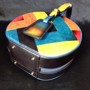 May include: A circular leather handbag with a colourful patchwork design. The top is patterned with red, orange, yellow, and teal panels. The bag has a black base with a blue zip and trim, and a matching luggage tag.