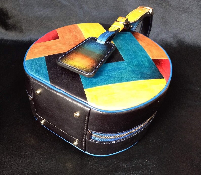 May include: A circular leather handbag with a colourful patchwork design. The top is patterned with red, orange, yellow, and teal panels. The bag has a black base with a blue zip and trim, and a matching luggage tag.