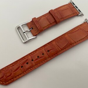 May include: A brown leather Apple Watch band with a silver buckle. The band has a crocodile pattern and is stitched with a yellow thread.