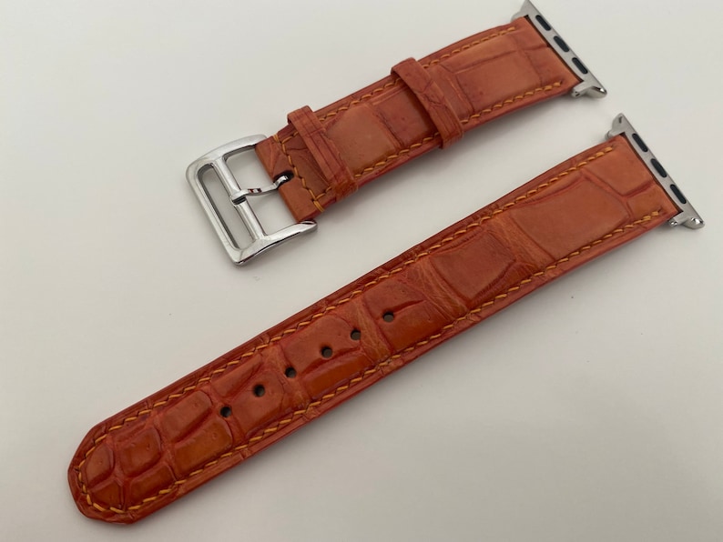 May include: A brown leather Apple Watch band with a silver buckle. The band has a crocodile pattern and is stitched with a yellow thread.