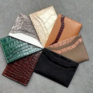Peut inclure: Une collection de porte-cartes en cuir de différentes couleurs et textures, dont vert, marron, noir et beige. Les porte-cartes présentent des motifs en relief, certains avec un design en peau de crocodile ou de serpent.