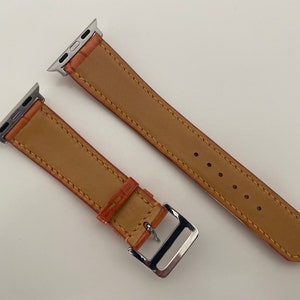 May include: Two brown leather Apple Watch bands with silver clasps. The bands have a stitched edge and are shown on a white background.
