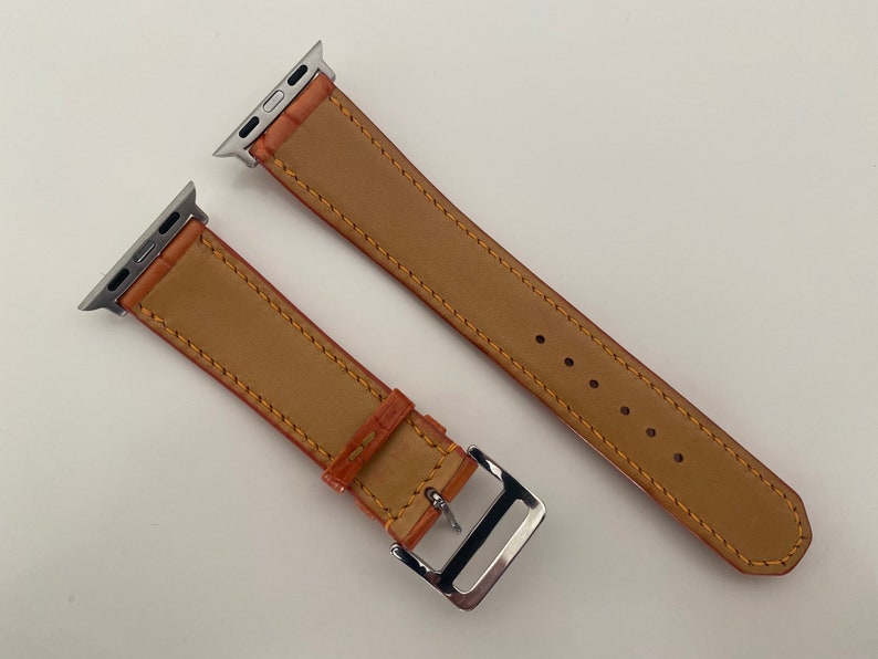 May include: Two brown leather Apple Watch bands with silver clasps. The bands have a stitched edge and are shown on a white background.