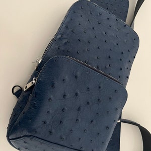 Op de afbeelding: Een donkerblauwe leren schoudertas met een textuur van struisvogelhuid. De tas heeft een ritssluiting en een verstelbare schouderband.